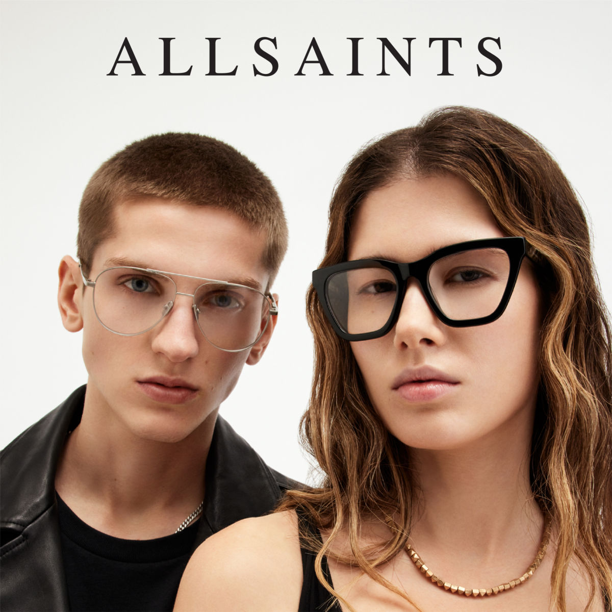 Two models stand against a white background, facing the camera. The short haired model on the left poses wearing aviator frames, the long haired model on the right poses wearing thick black angular frames. AllSaints logo is displayed across the top of the image.