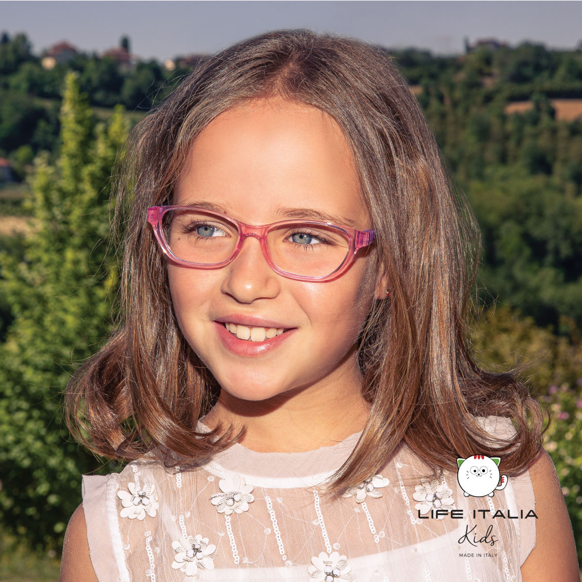 Young girl with shoulder length brown hair, standing outside, wearing pink translucent frames. LIFE ITALIA KIDS logo imposed on lower right of image.