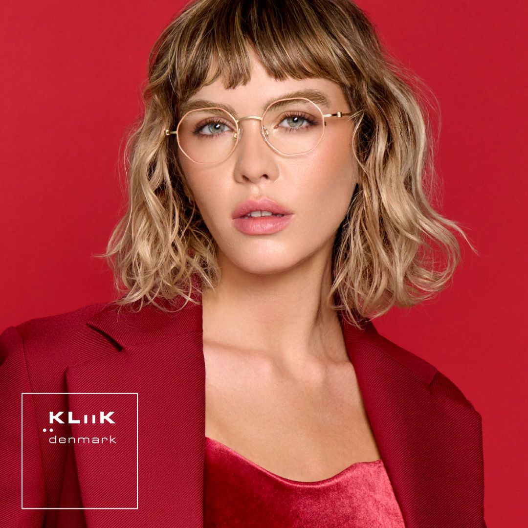 Woman with shoulder length wavy blonde hair wearing a red sport coat over a shiny red shirt against a red background, models delicate geometric blush and gold frames. KLiiK denmark logo in a white box on the bottom left of the image.