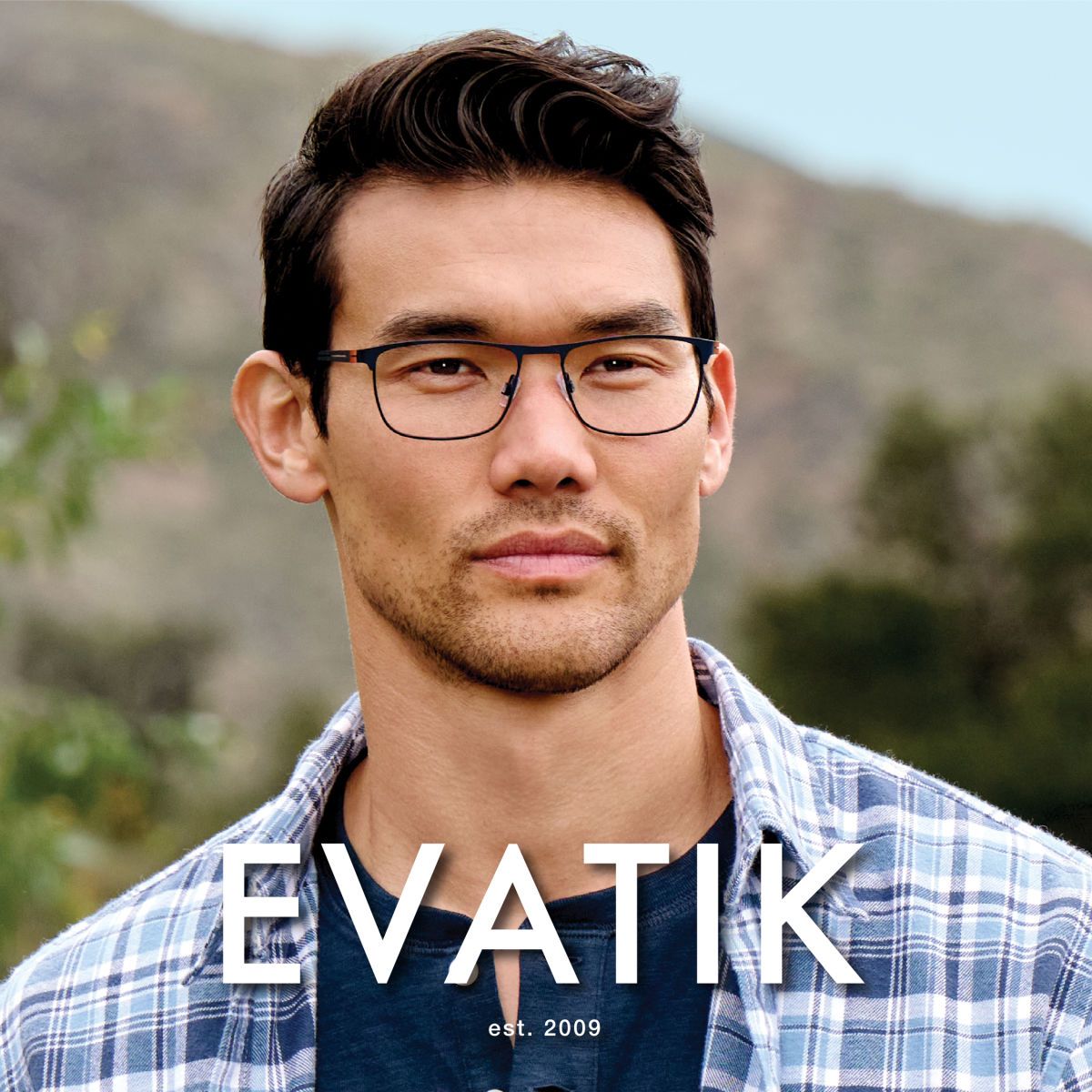 Man with dark wavy hair wearing a blue plaid shirt, faces to the right of the camera to show off square frames. EVATIK logo in white along the bottom.