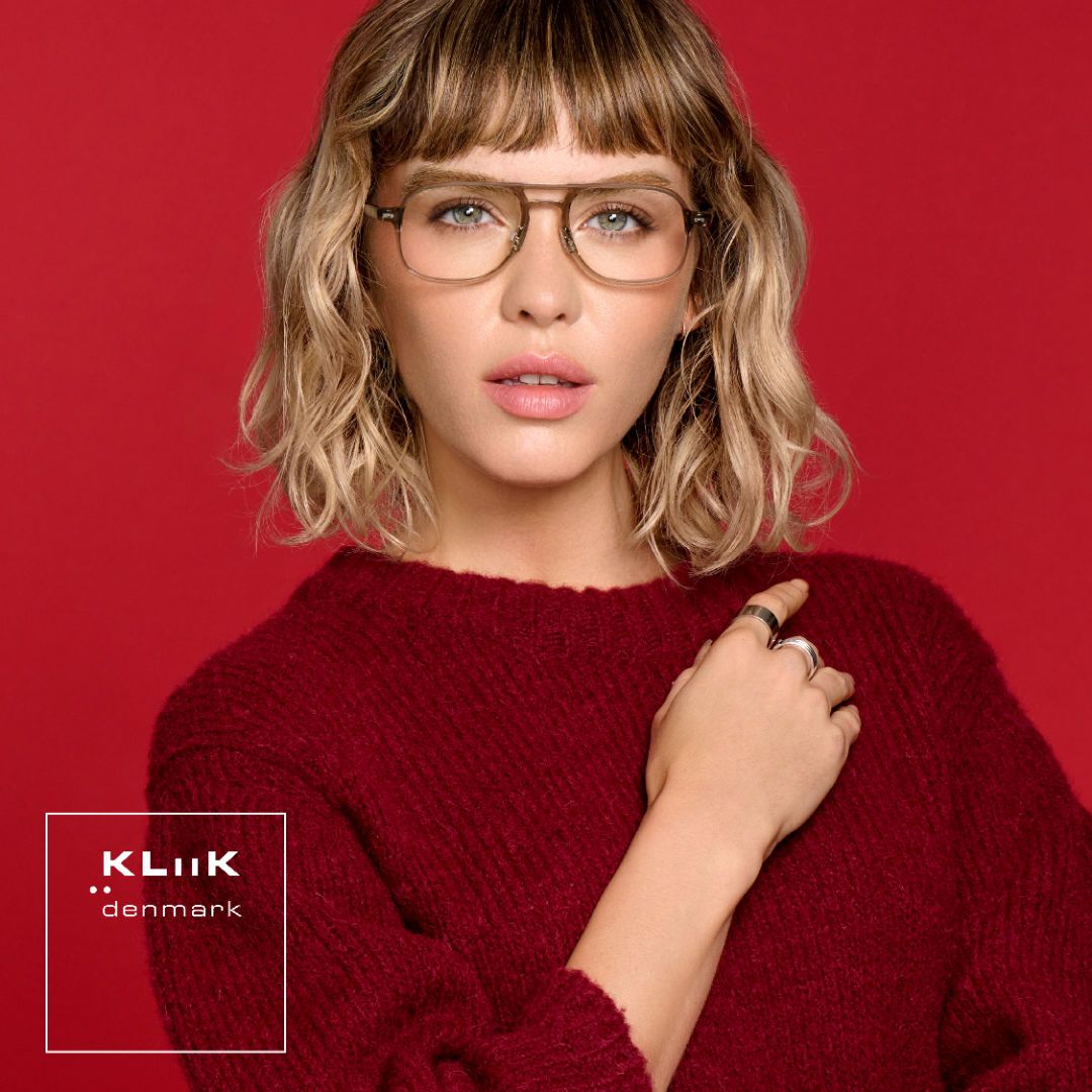 Woman with shoulder length wavy blonde hair wearing a dark red sweater against a red background, models gold modified aviator frames. KLiiK denmark logo in a white box on the bottom left of the image.
