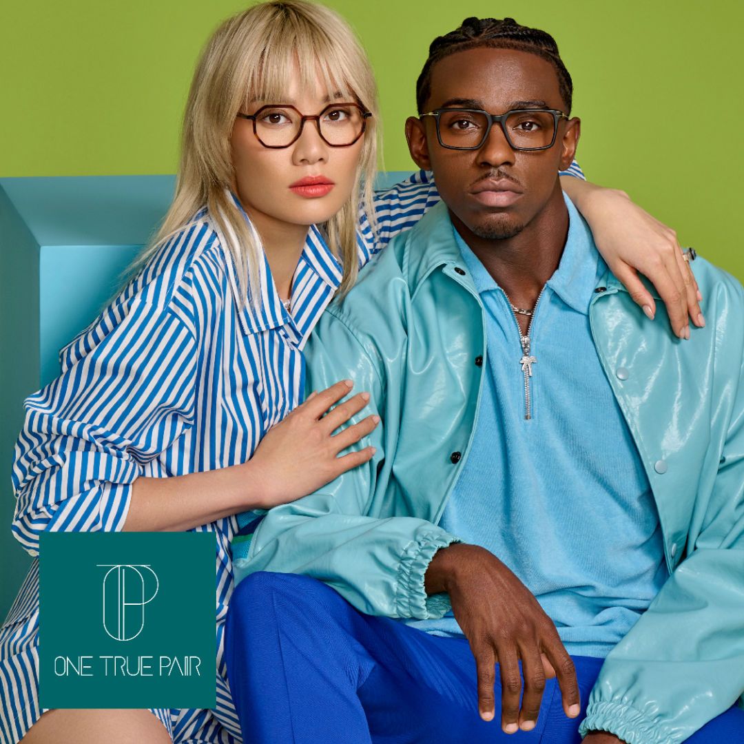 An Asian woman with blonde hair and a blue and white stripped shirt poses with her arm around the shoulders of a Black man wearing a light blue shirt and jacket. The woman is on the left and models a pair of thick octegonal frames, the man is on the right modeling thick dark frames with gold accents. One True Pair logo located bottom left.