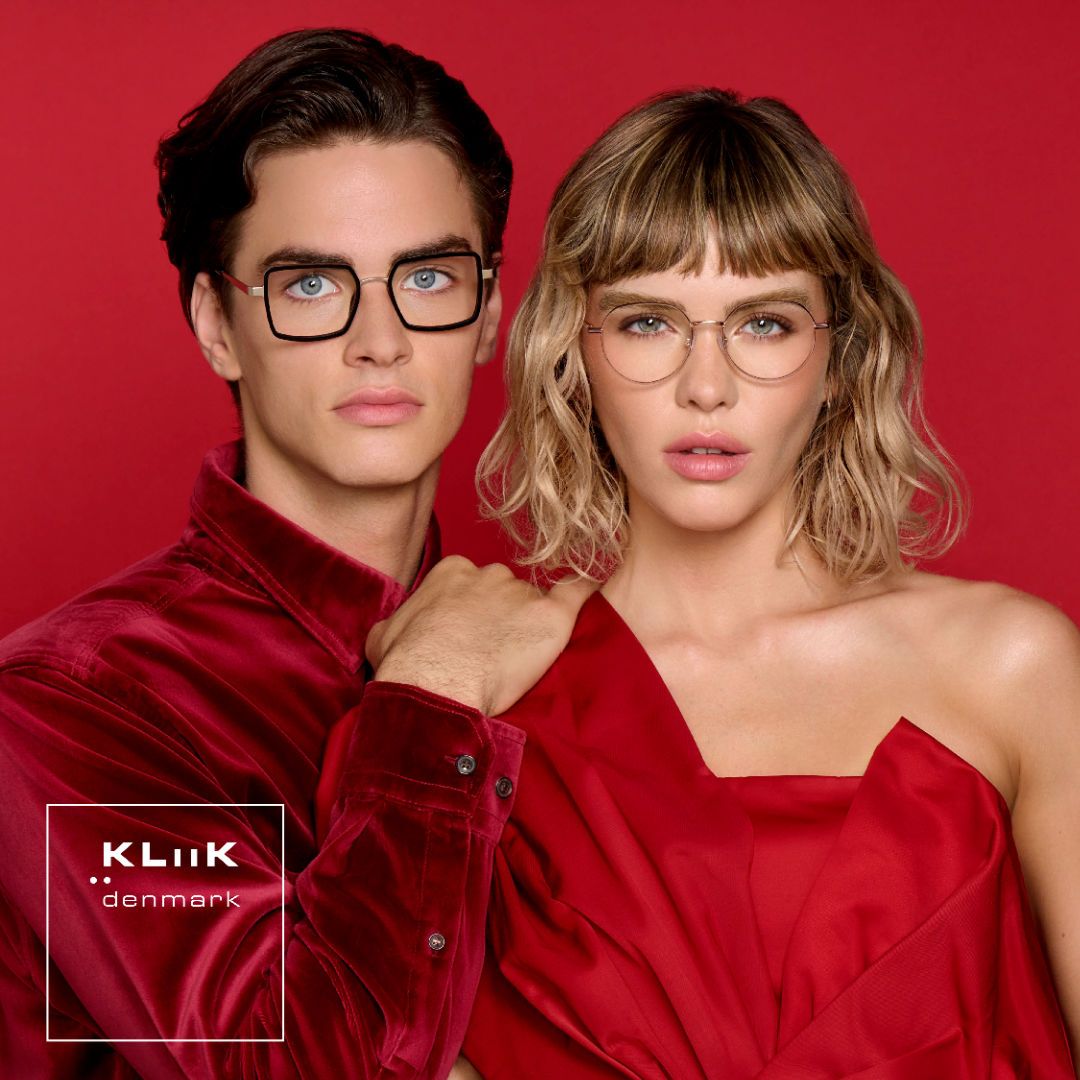 Two models stand against a red background facing the camera. On the left is a man wearing a red velor collared shirt modeling thick dark square frames. On the right a is a woman with shoulder length wavy blond hair wearing a red one-shouldered dress modeling delicate round gold frames. KLiiK denmark logo in a white box on the bottom left of the image.