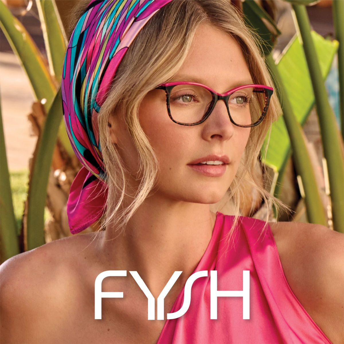 A close-up of a blonde model wearing a bright colorful head scarf, stylish frames that are mostly black with a pop of pink along the top rim. The Fysh logo is displayed in white across the bottom center of the image.