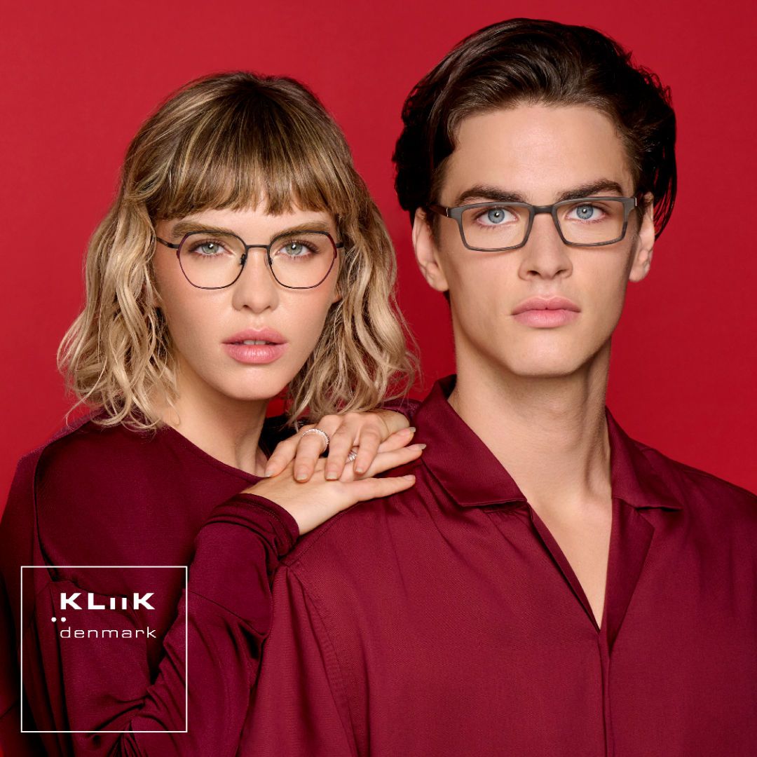 Two models stand against a red background facing the camera. On the left is a woman with shoulder length wavy blonde hair, wearing a dark red long-sleeved shirt, modeling delicate dark geometric frames. On the right a is a man with short brown hair, wearing a red collared shirt, modeling thick dark rectangular frames. KLiiK denmark logo in a white box on the bottom left of the image.
