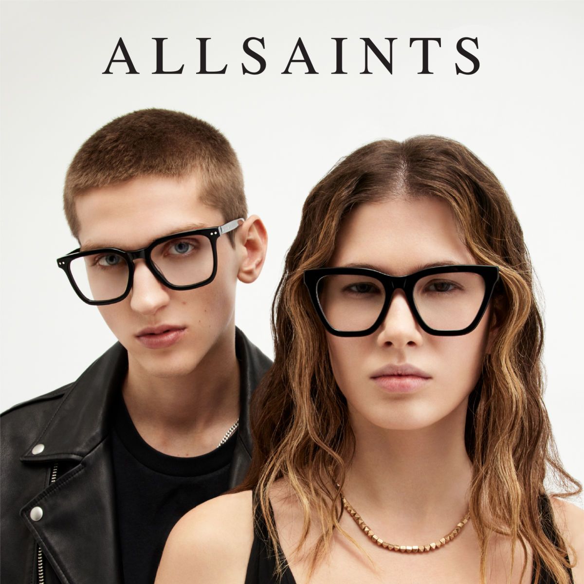 Two models stand against a white background, facing the camera, each wearing a different style thick black AllSaints frames. AllSaints logo is displayed across the top of the image.