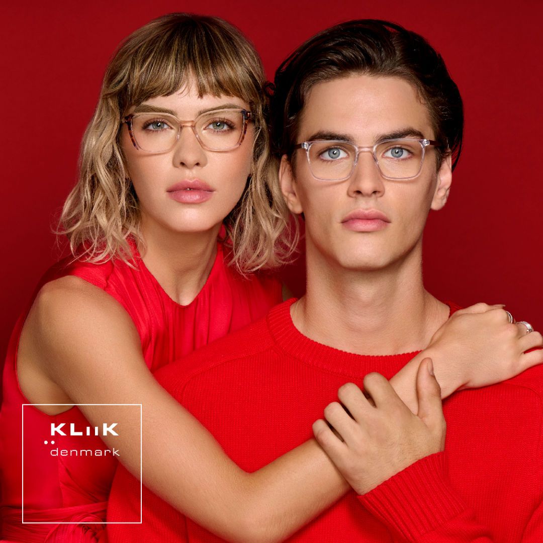 Two models stand against a red background facing the camera. On the left is a woman with shoulder length wavy blonde hair, wearing a bright red sleeveless shirt, modeling bold two-toned modified cat-eye frames. On the right a is a man with short brown hair, wearing a bright red sweater, modeling thick clear square frames. KLiiK denmark logo in a white box on the bottom left of the image.