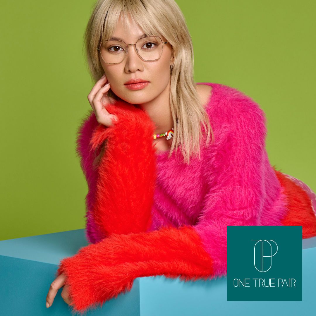 An Asian woman in a fuzzy pink and orange sweater poses againts a green background modeling a pair of thin gold frames.