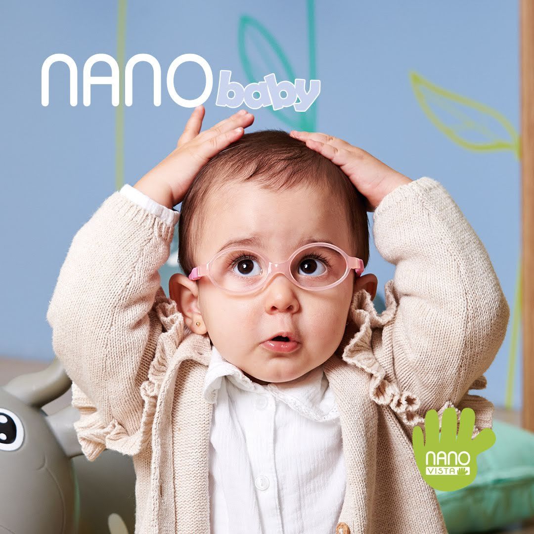 Young baby in a white knit sweater sitting on the floor with her hands on her head weating light pink frames straped to her head.. Nano Baby logo located in the upper left, Nano Vista logo located on bottom right.