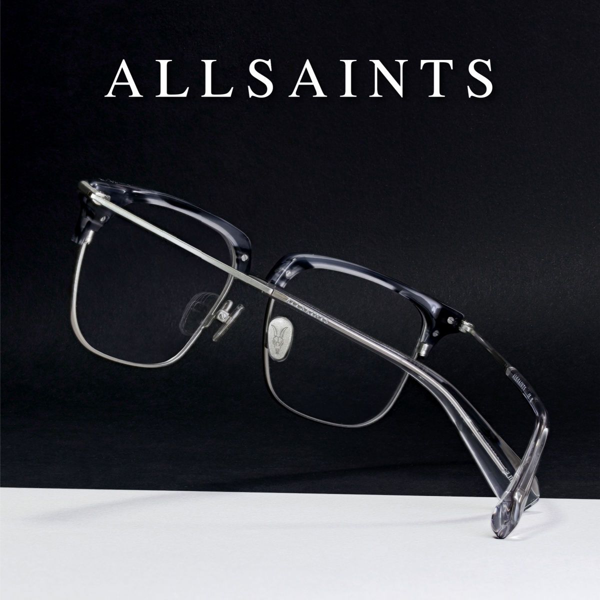 Frames with a thick black top and thin wire lower frame on a black and white background, facing away to show the crossed temples. AllSaints logo in white at the top of the image.