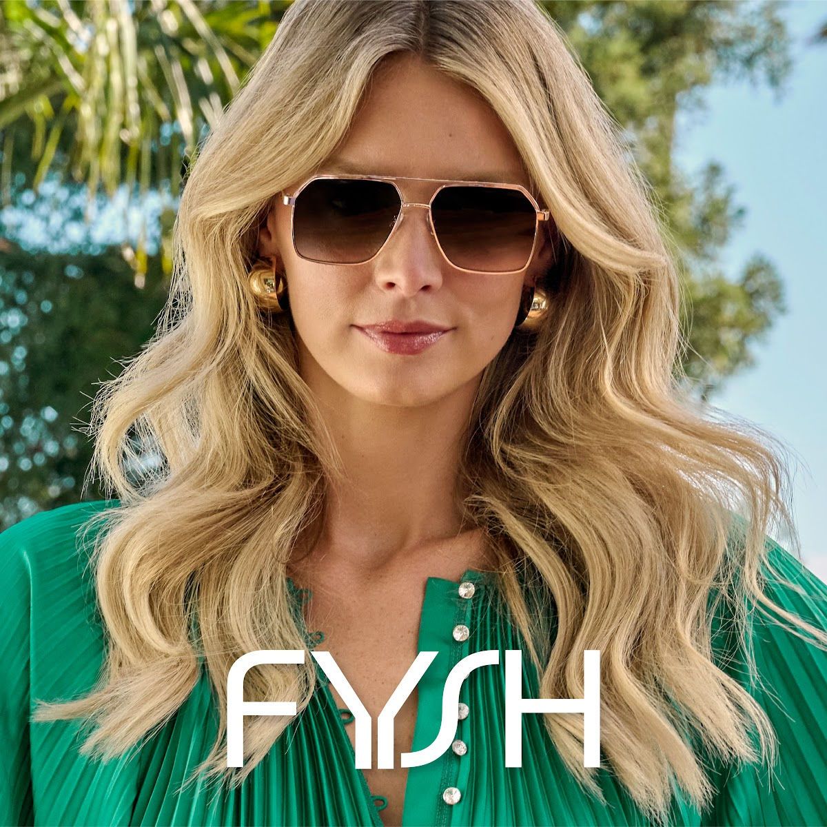 A woman with long wavy blonde hair modeling rose-gold aviator style sunglasses, wearing a bright green pleated shirt, and standing outside with trees and blue sky in the background. The Fysh logo is displayed in white across the bottom center of the image.