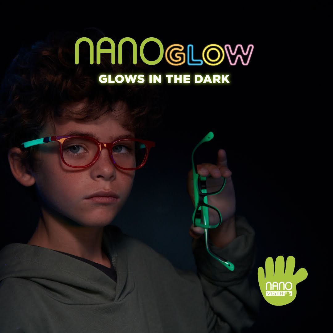 Young boy with curly hair, a grey hoodie and a serious expression stands to the left against a dark backround, wearing red frames with glowing green temples, while holding and bending a different set of frames. Nano Glow Glows in the dark is written across the top of the image, Nano Vista logo located on bottom right.