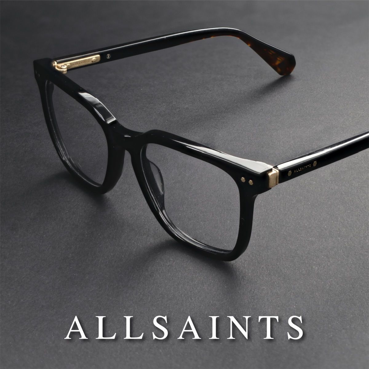 Thick black frames sitting propped open, facing lower left, on a grey surface. White AllSaints logo runs along the bottom of the image.