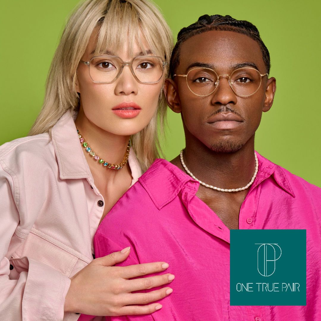 An Asian woman with blonde hair and a light pink shirt poses with her hand on the arm of a Black man in a bright pink shirt against a bright green background. The woman is on the left and is modeling thick clear frames, the man is on the right and is modeling thin gold frames. One True Pair logo located bottom right. 