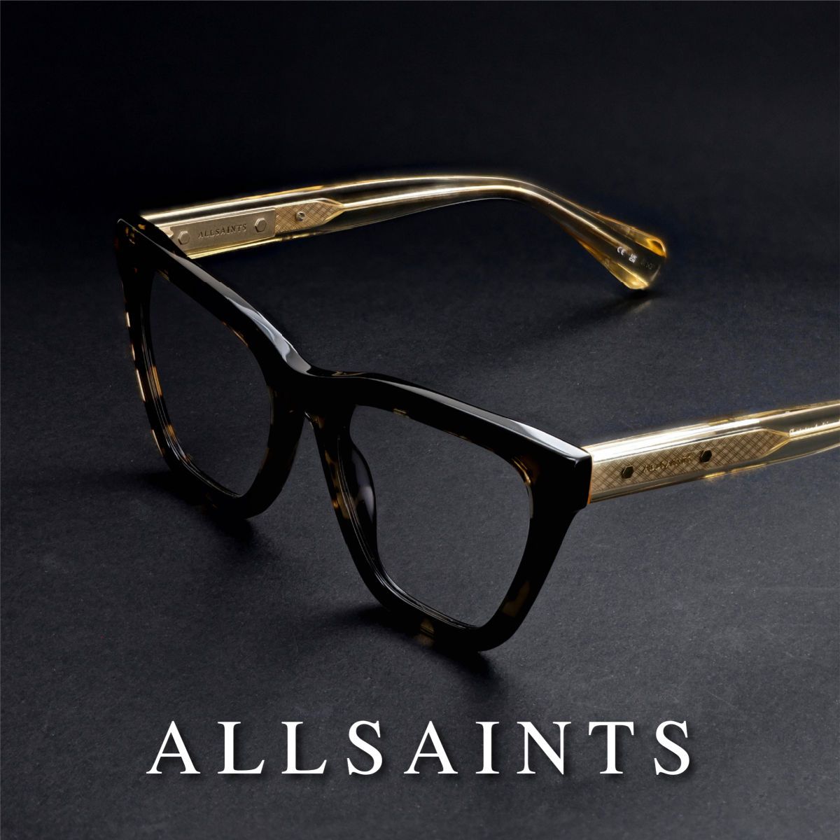 Thick black frames sitting propped open showing gold temples, facing lower left, on a grey surface. White AllSaints logo runs along the bottom of the image.