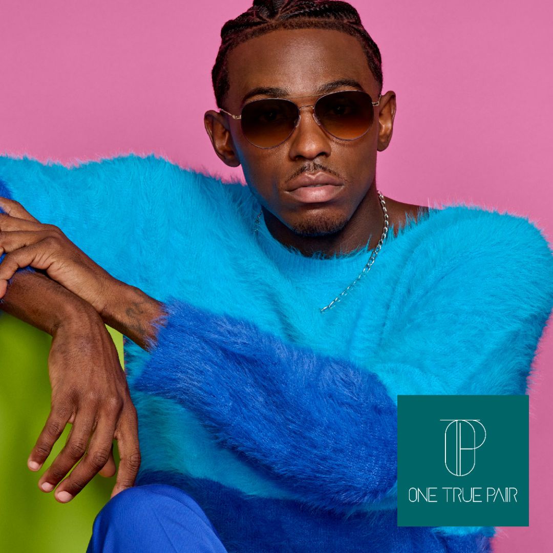 A black man in a fuzzy blue sweater sits in a casual pose against a pink backgroung modeling aviator style sunglasses. One True Pair logo located bottom right.