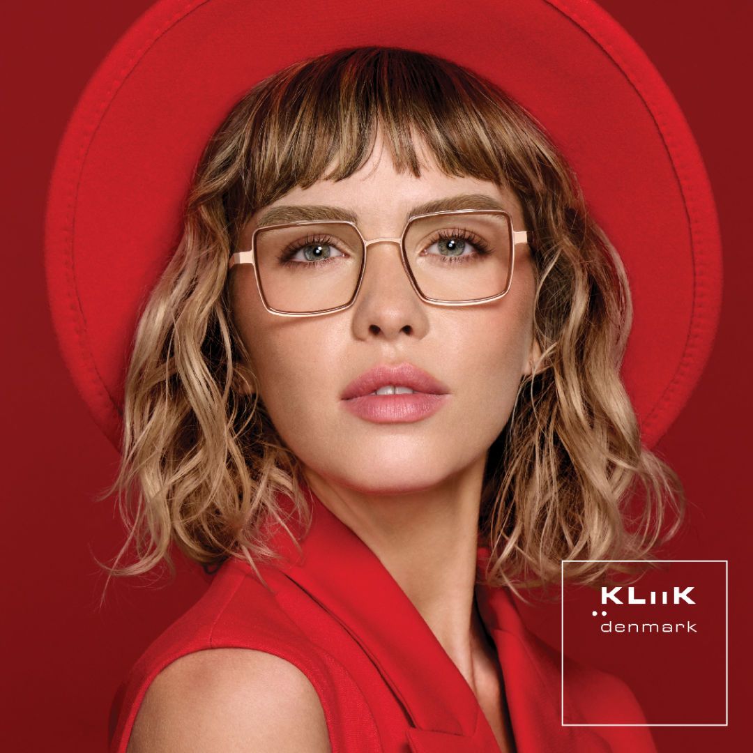 Woman with shoulder length wavy blonde hair wearing a red collared sleeveless shirt and red round brimmed hat against a red background, models bold square gold frames. KLiiK denmark logo in a white box on the bottom right of the image.