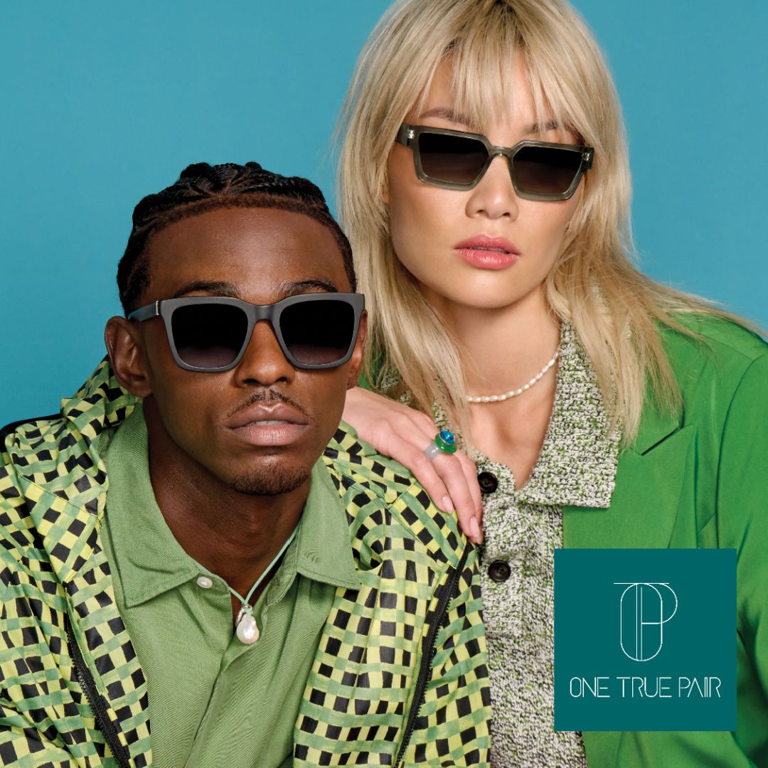 An Asian woman with blonde hair and a green shirt poses with her arm on the shoulder of a Black man wearing a green patterned jacket. The woman is on the right and models a pair of thick opaque square sunglasses, the man is on the left modeling thick dark square sunglasses. One True Pair logo located bottom right.
