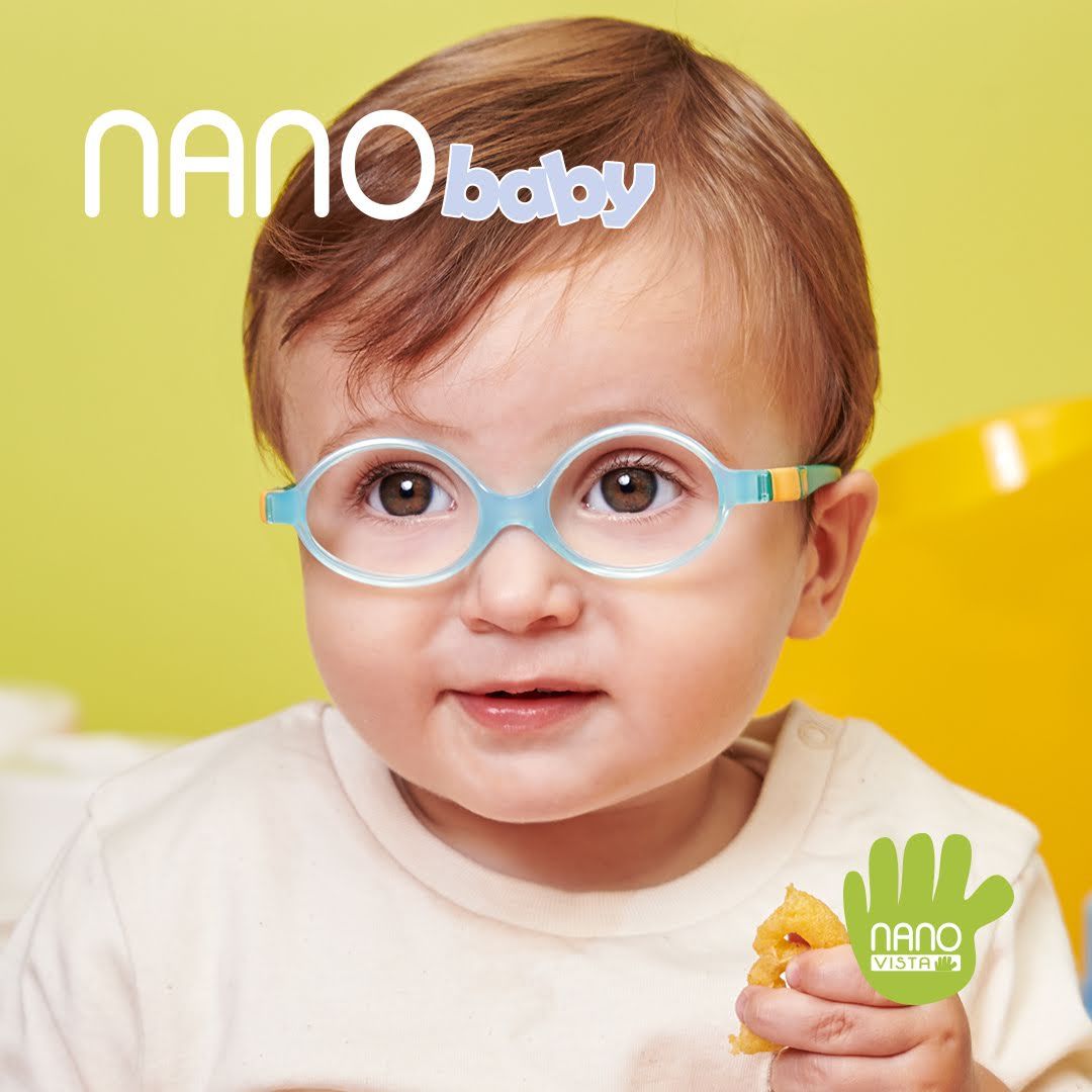 Young baby in a white onesie sitting against a yellow backgrounds weaing light ble frames with yellow and green temples, holding a half eaten snack in his hand. Nano Baby logo located in the upper left, Nano Vista logo located on bottom right.