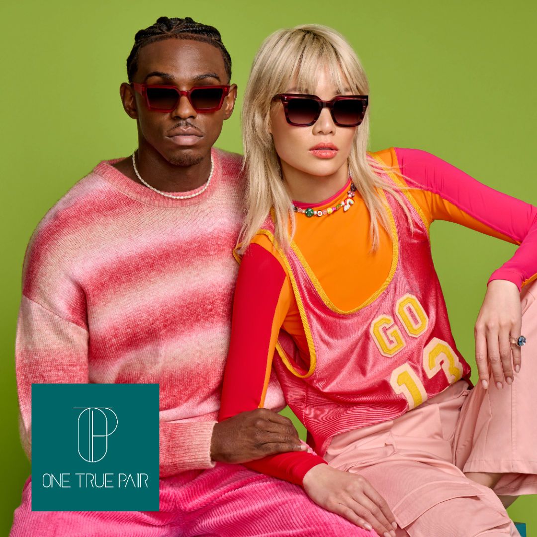 A black man in a pink striped sweater modeling thick framed red sunglasses, sits with an asian woman with blond hair wearing a pink jersey modeling thick framed opaque red square sunglasses. One True Pair logo located on bottom left.