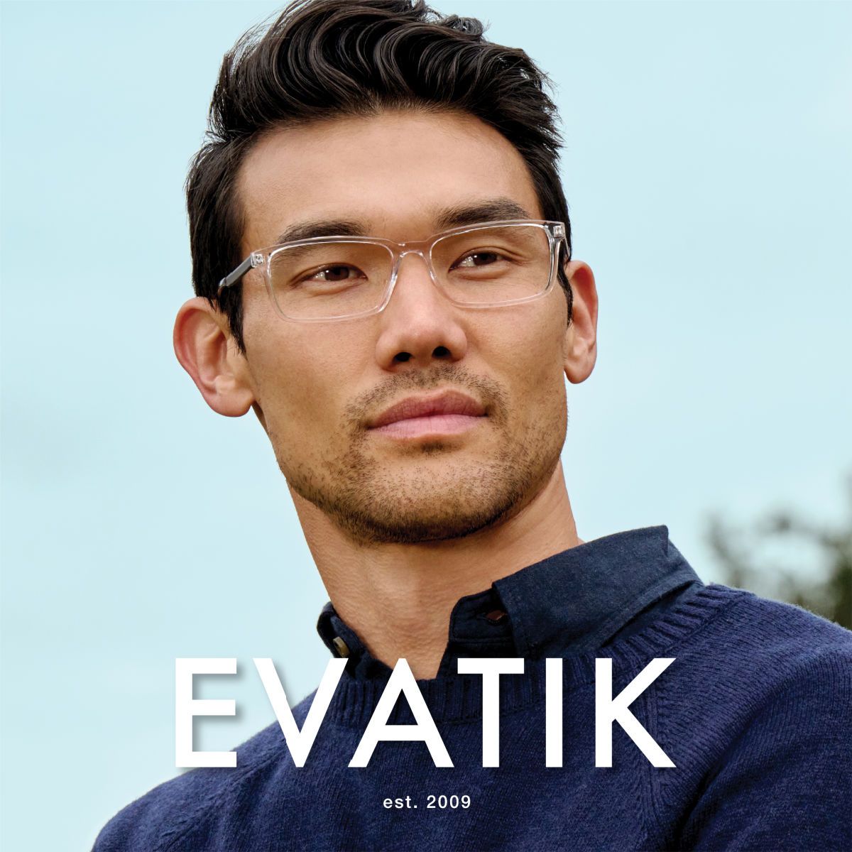 Man with dark wavy hair and a dark blue sweater stands against a bright blue sky, faces off to the right of the camera to show off transparent square frames with black temples. EVATIK logo in white along the bottom.