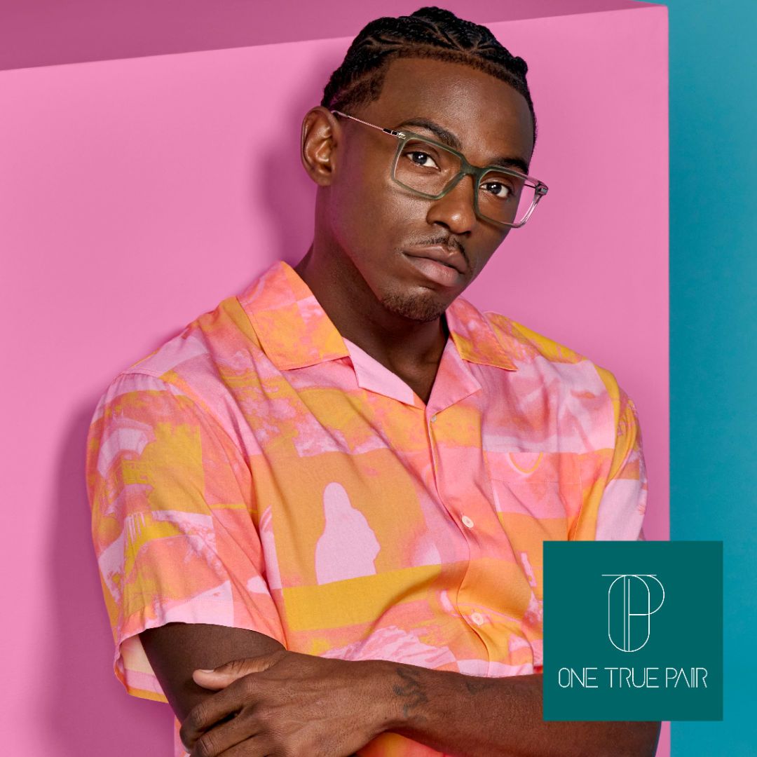 A black man wearing a pink and orange button-up short sleeved shirt, with arms folded, leaning against a pink and blue background, modeling thick square frames with slender temples. One True Pair logo at bottom right.
