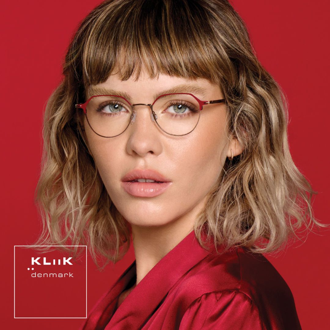 Woman with shoulder length wavy blonde hair wearing a shiny red shirt against a red background, models delicate geometric red and gold frames. KLiiK denmark logo in a white box on the bottom left of the image.