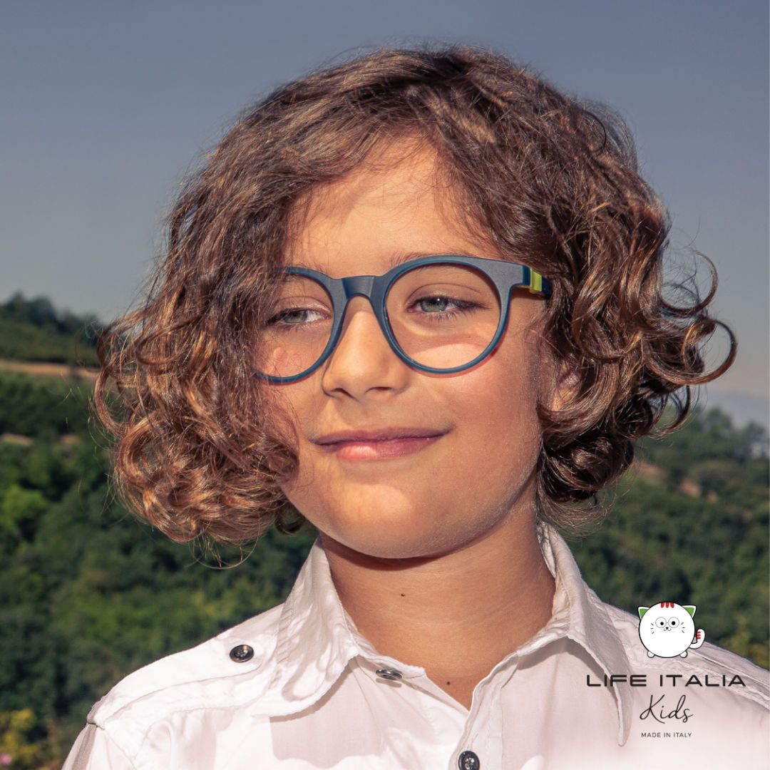 Curly haired kid standing outside wearing thick round dark blue frames with a hint of green on the temple. LIFE ITALIA KIDS logo imposed on lower right of image.