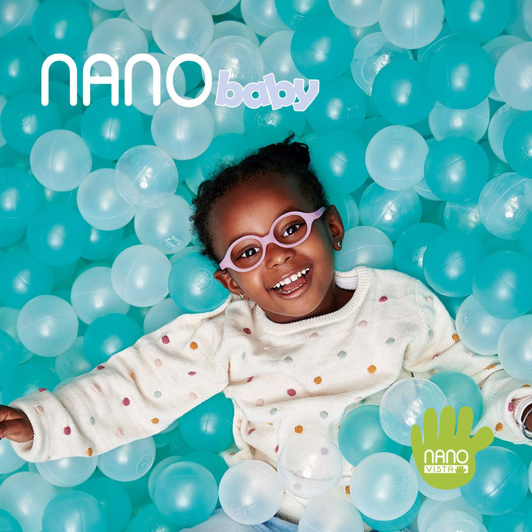 African American baby in a white polka dot sweater and bright pink round frames, grinning while laying in a ball pit of blue and clear balls. Nano Baby logo located in the upper left, Nano Vista logo located on bottom right.