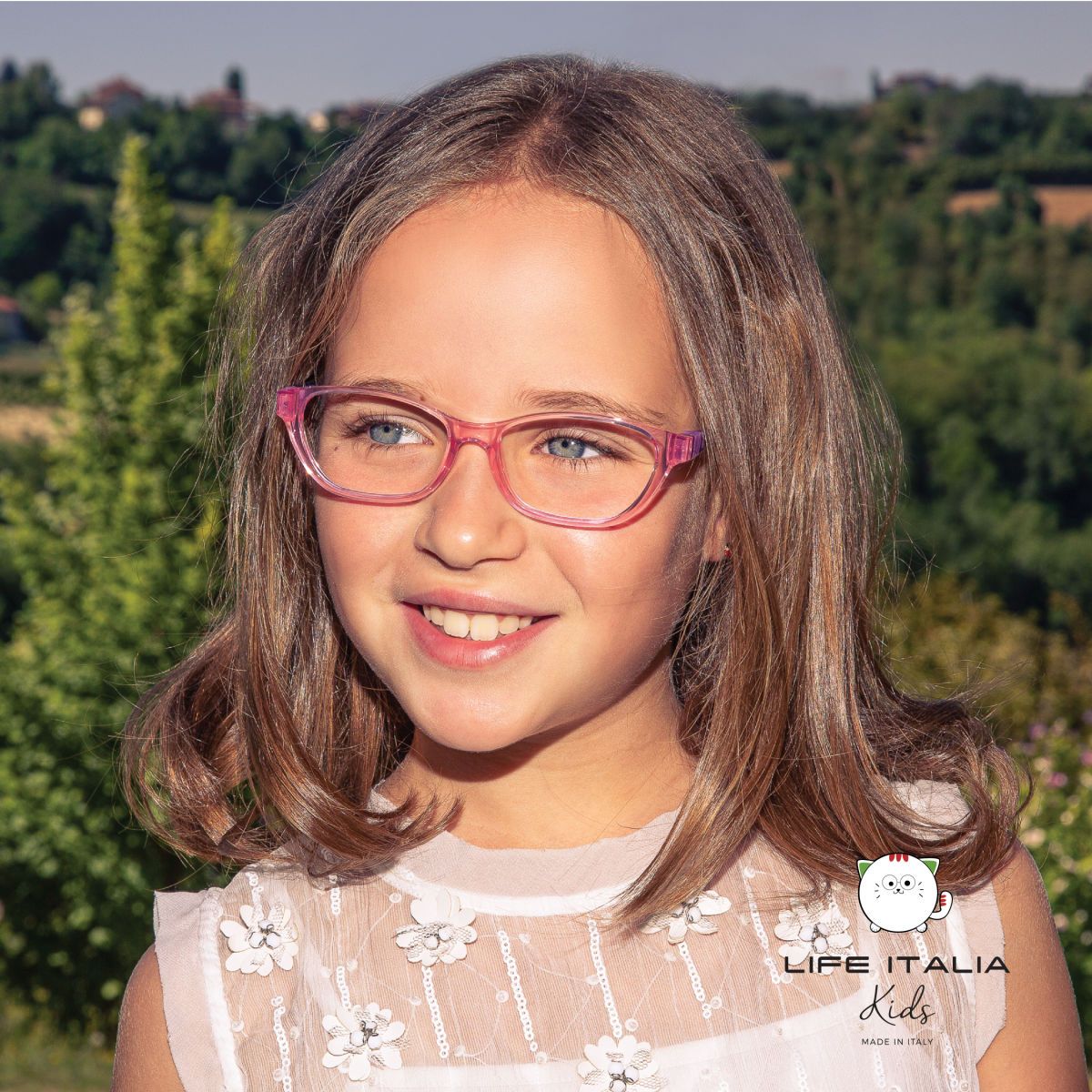 Young girl with shoulder length brown hair, standing outside, wearing pink translucent frames. LIFE ITALIA KIDS logo imposed on lower right of image.