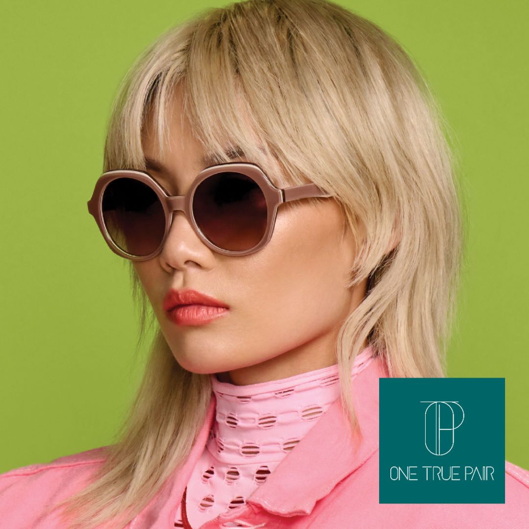 An asian woman with blong hair wearing a fashionable pink top stands against a green background facing off to the left, modeling a pair of thick tan round sunglasses. One True Pair logo located bottom right.