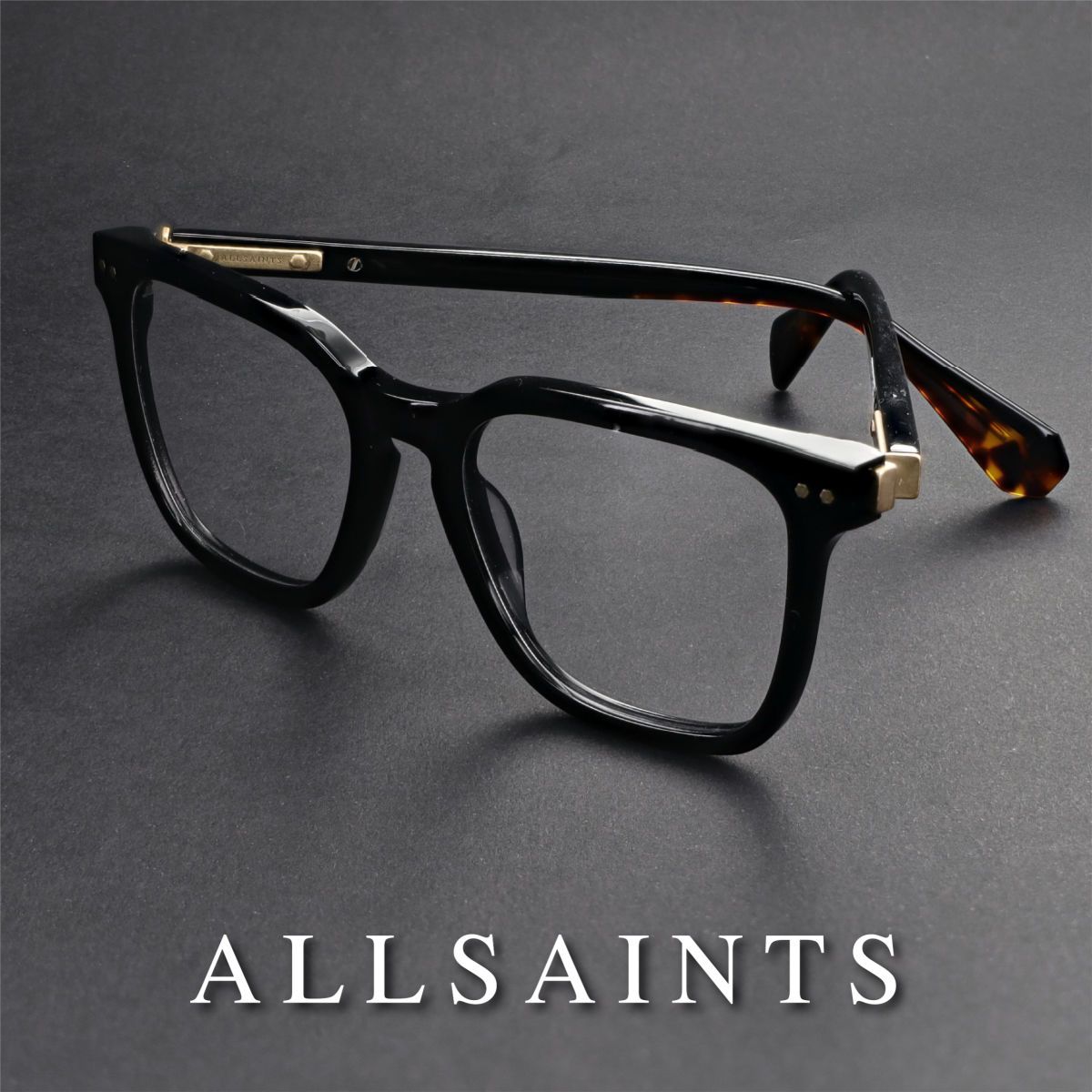 Thick black frames propped open with temples crossed, facing lower right, on a grey background. AllSaints logo in white text along the bottom of the image.
