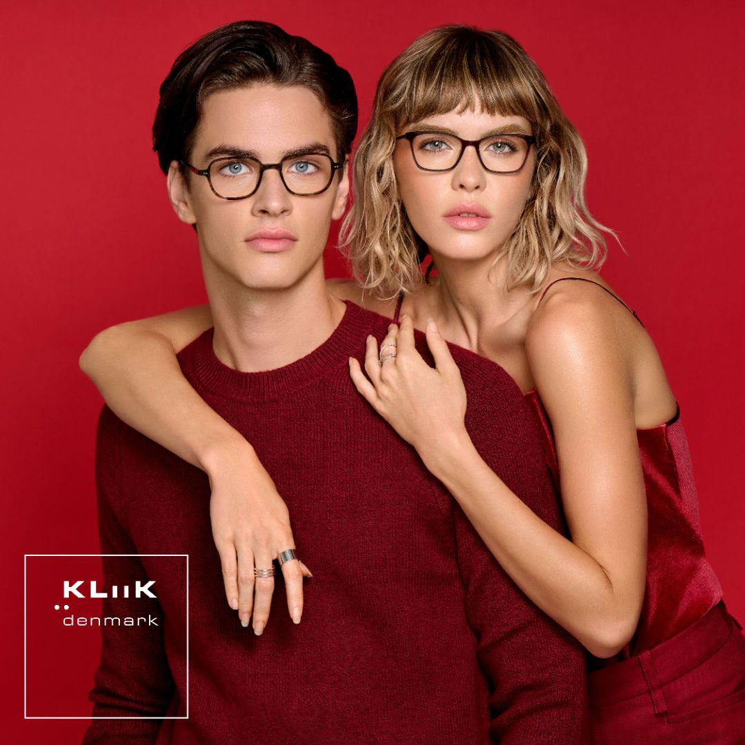 Two models pose against a red background facing the camera. On the left is a man wearing a dark red sweater is modeling thick dark rounded frames. On the right is a woman with shoulder length wavy blond hair, arms draped around the male model, wearing a red thin-strapped shirt, modeling thick dark modified cat-eye frames. KLiiK denmark logo in a white box on the bottom left of the image.