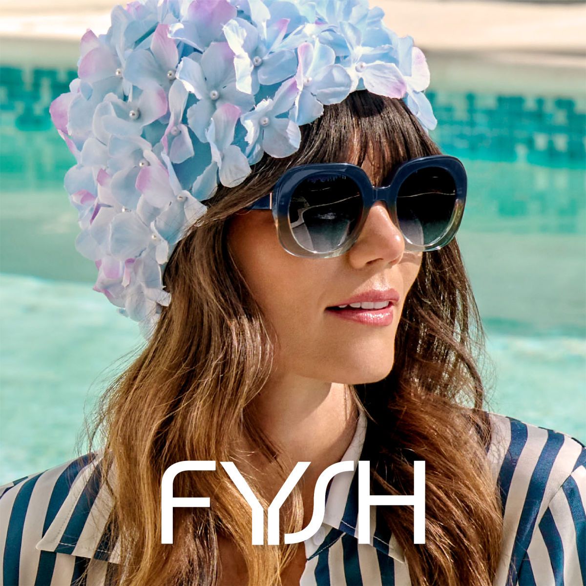 Close-up of a woman with long wavy brown hair in a faux flowery swim hat against a blurred outdoor pool, models two-toned blue to clear sunglasses. The Fysh logo is displayed in white across the bottom center of the image against the models blue and white striped shirt.