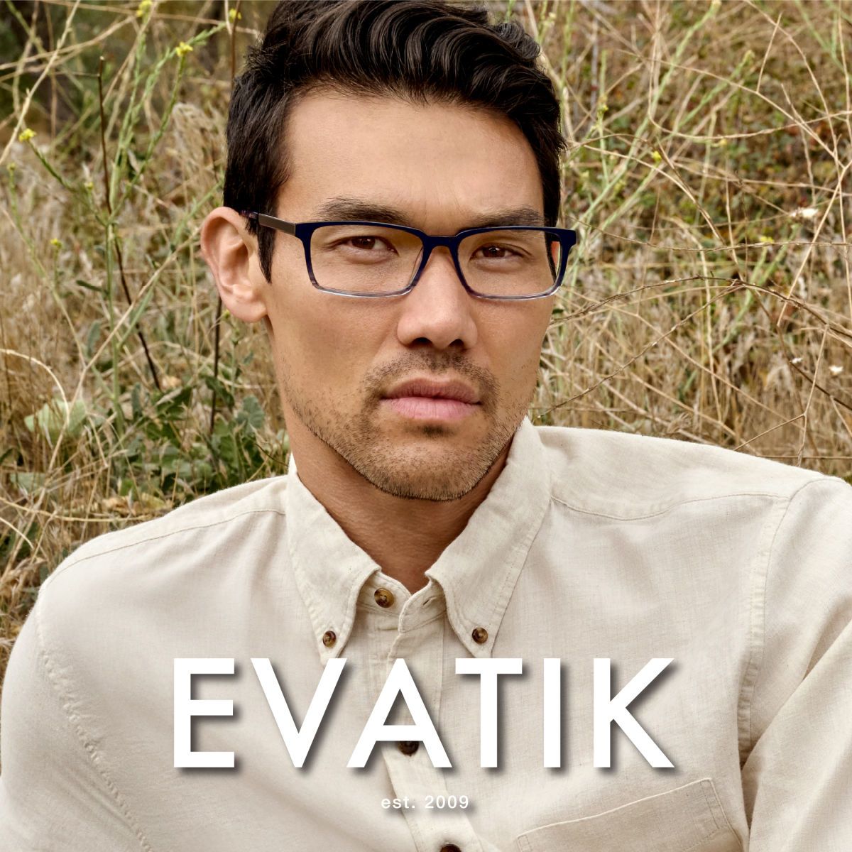 A man with dark wavy hair and white button up shirt, sits in tall grass facing the camera modeling square frames that are dark on the top and fade to transparent. EVATIK logo in white along the bottom.