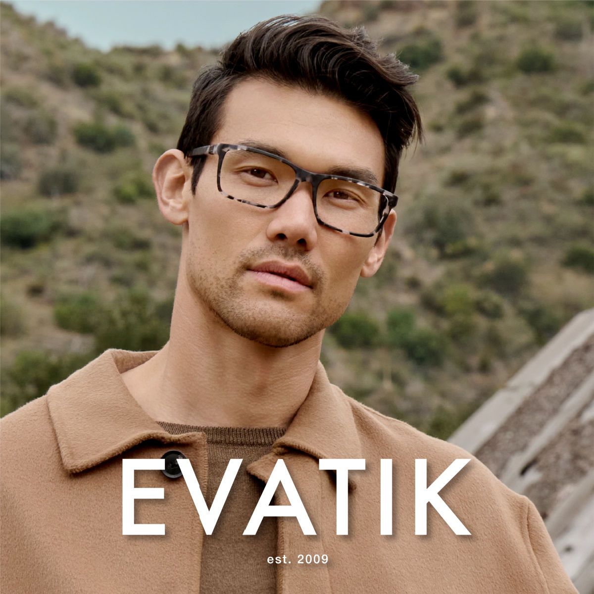 Man with dark wavy hair and a dark brown coat stands against a scrubby hillside, faces the camera to show off matt grey tortoise patterned square frames. EVATIK logo in white along the bottom.