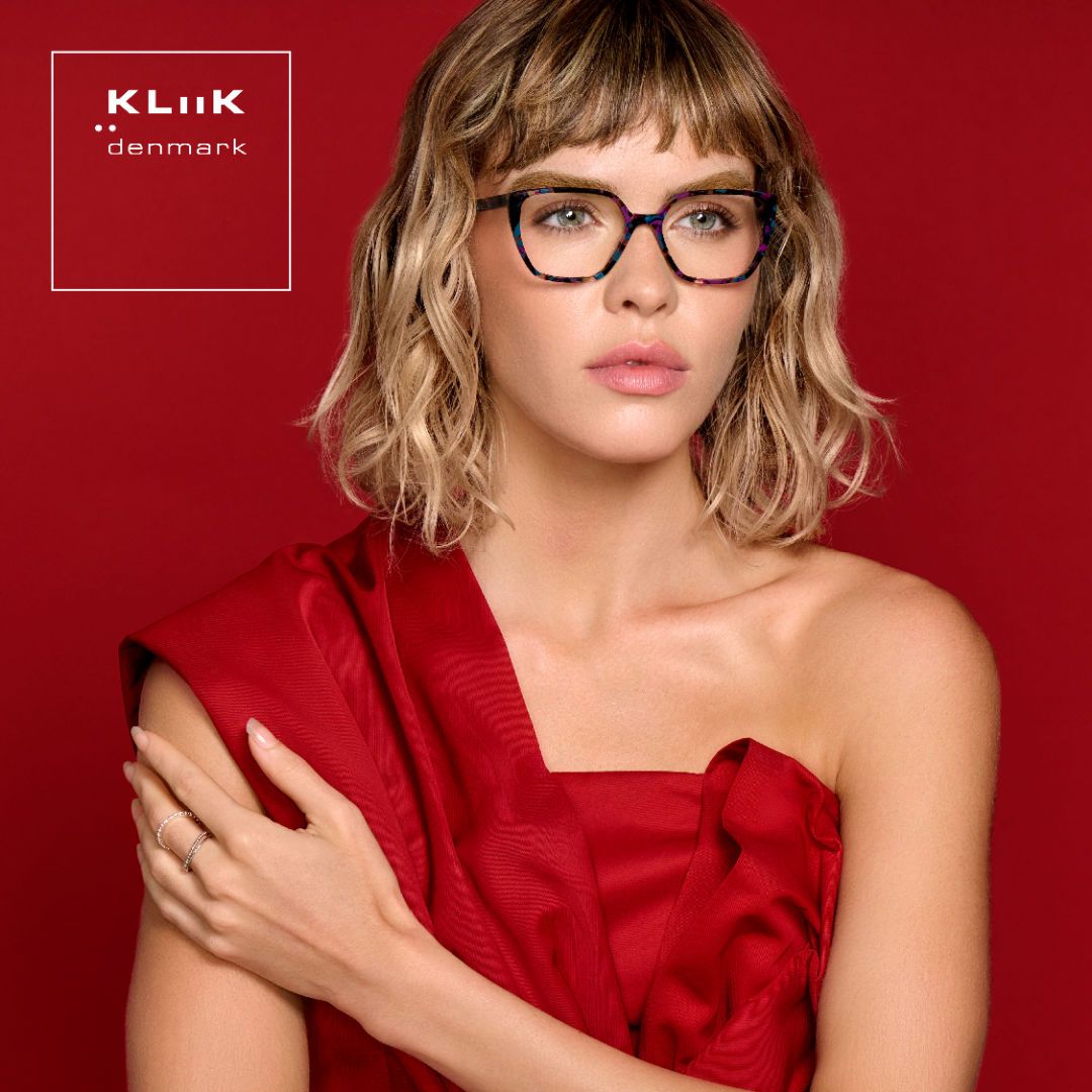Woman with shoulder length wavy blonde hair wearing a one-shouldered red dress stands against a red background, modeling an acetate frame featuring a stylish black confetti print. KLiiK denmark logo in a white box on the top left of the image.