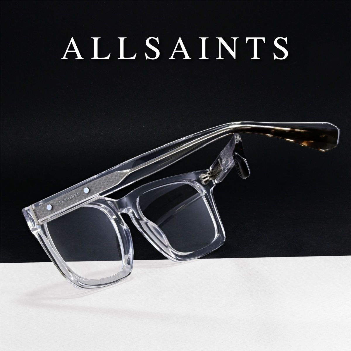 Thick clear frames on a black and white background, facing away to show the crossed temples. AllSaints logo in white at the top of the image.