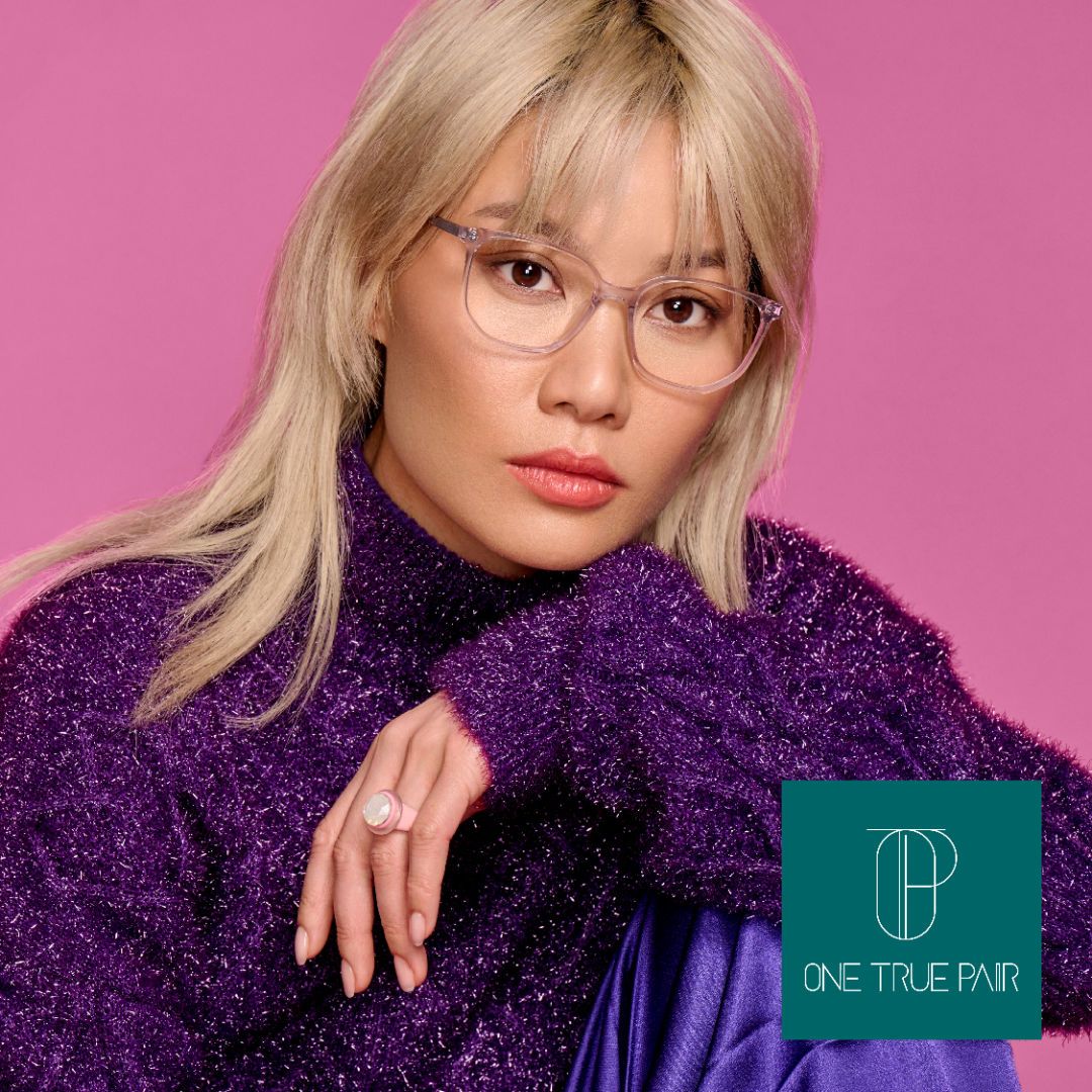 An Asian woman with blonde hair and a fuzzy purple sparkly sweater poses against a pink background modeling thick translucent frames. One True Pair logo located bottome right.