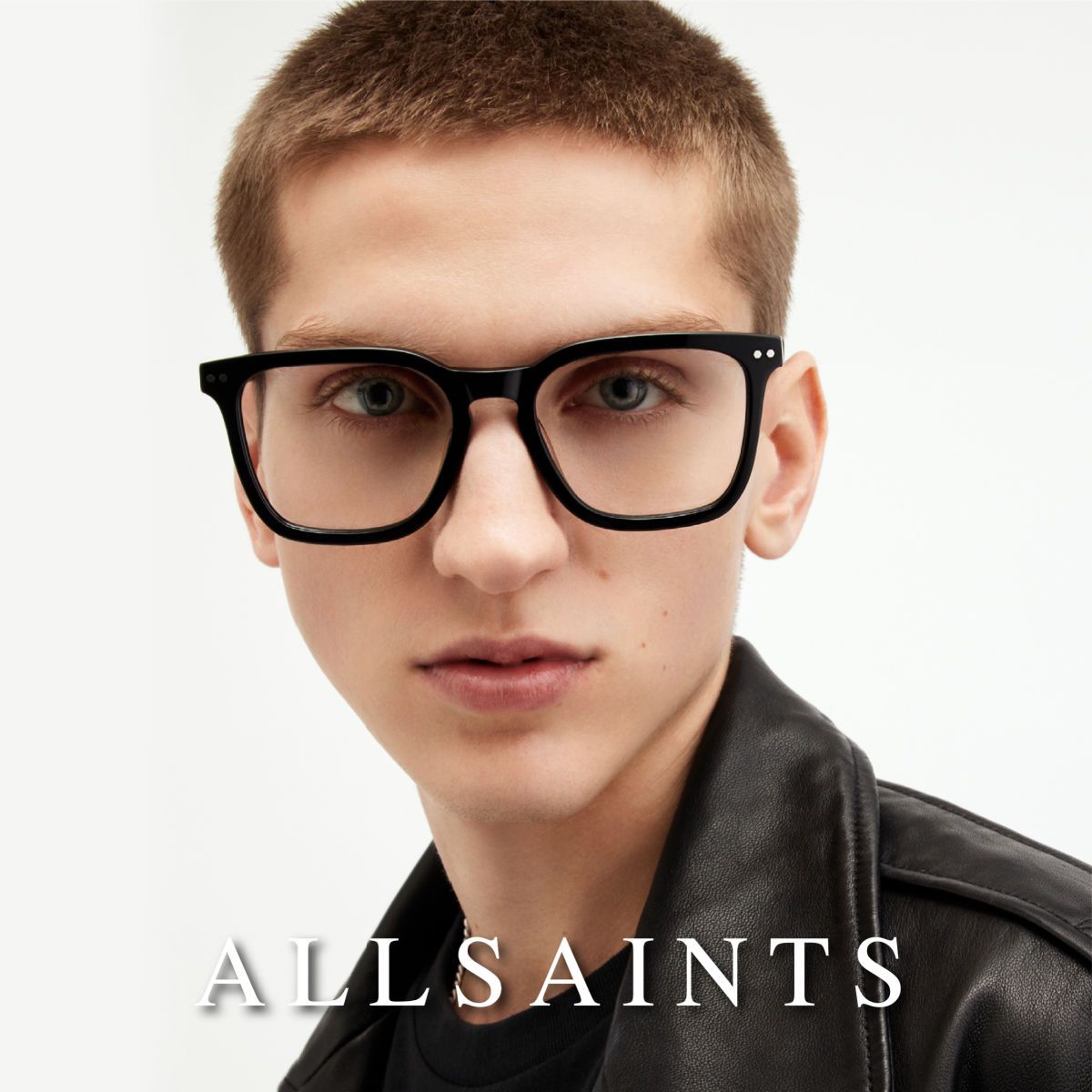 A model with short hair, black leather jacket, and thick black frames facing camera, against a white background. AllSaints logo in white along the bottom