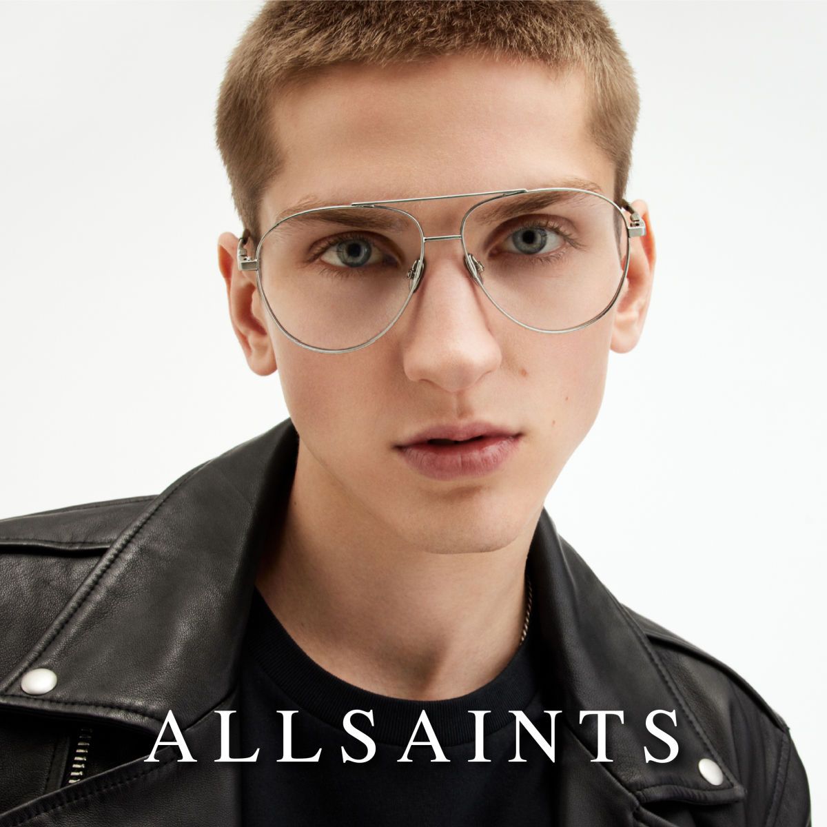 A model with short hair, black leather jacket, and double bridge aviator frames facing camera, against a white background. AllSaints logo in white along the bottom.