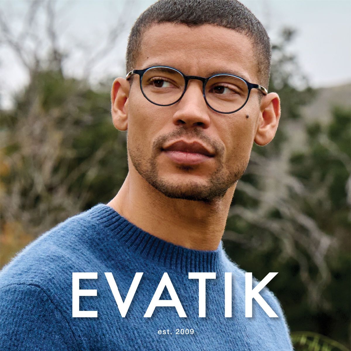 Man with short black hair in a blue sweater stands in front of scrub trees, facing slightly left of camera showing off round frames. EVATIK logo in white along the bottom.